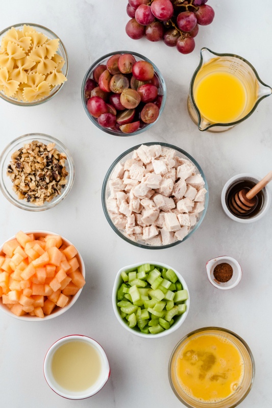 Fresh Fruity Chicken Pasta Salad - Ingredients in glass bowls - bow tie pasta, grapes, orange juice, beaten eggs, honey, nutmeg, cubed cantaloupe, cooked cubed chicken, sliced celery, lemon juice, and chopped walnuts