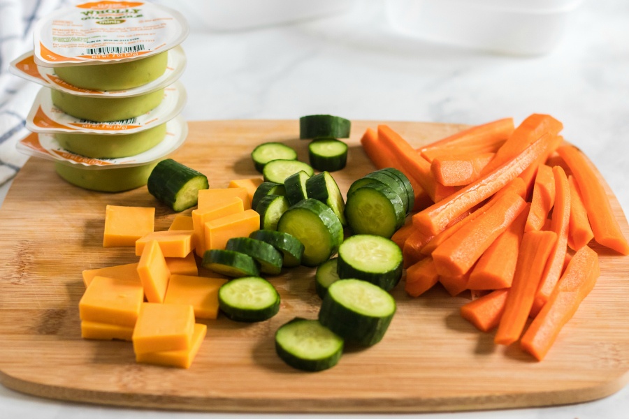  lunchbox ingredients on a cutting board - cheese, guacamole, cucumbers and carrots